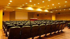 Un amplio auditorio interior con hileras de sillas verdes y un escenario con un atril rojo.
