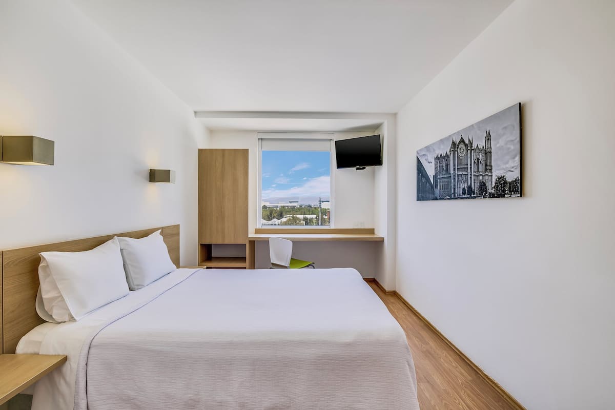 A hotel room with a bed, a desk, a chair, a TV, and a painting on the wall.