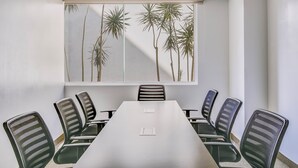 Un salón de conferencias con una larga mesa y seis sillas, un ventanal con vistas a palmeras, y un techo blanco.