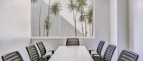 Une salle de conférence avec une longue table et six chaises, une grande fenêtre donnant sur des palmiers et un plafond blanc.