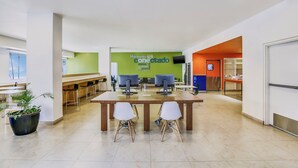 Un espacio interior moderno y luminoso con una mesa grande y sillas, una pared verde con texto, y una zona de cocina al fondo.