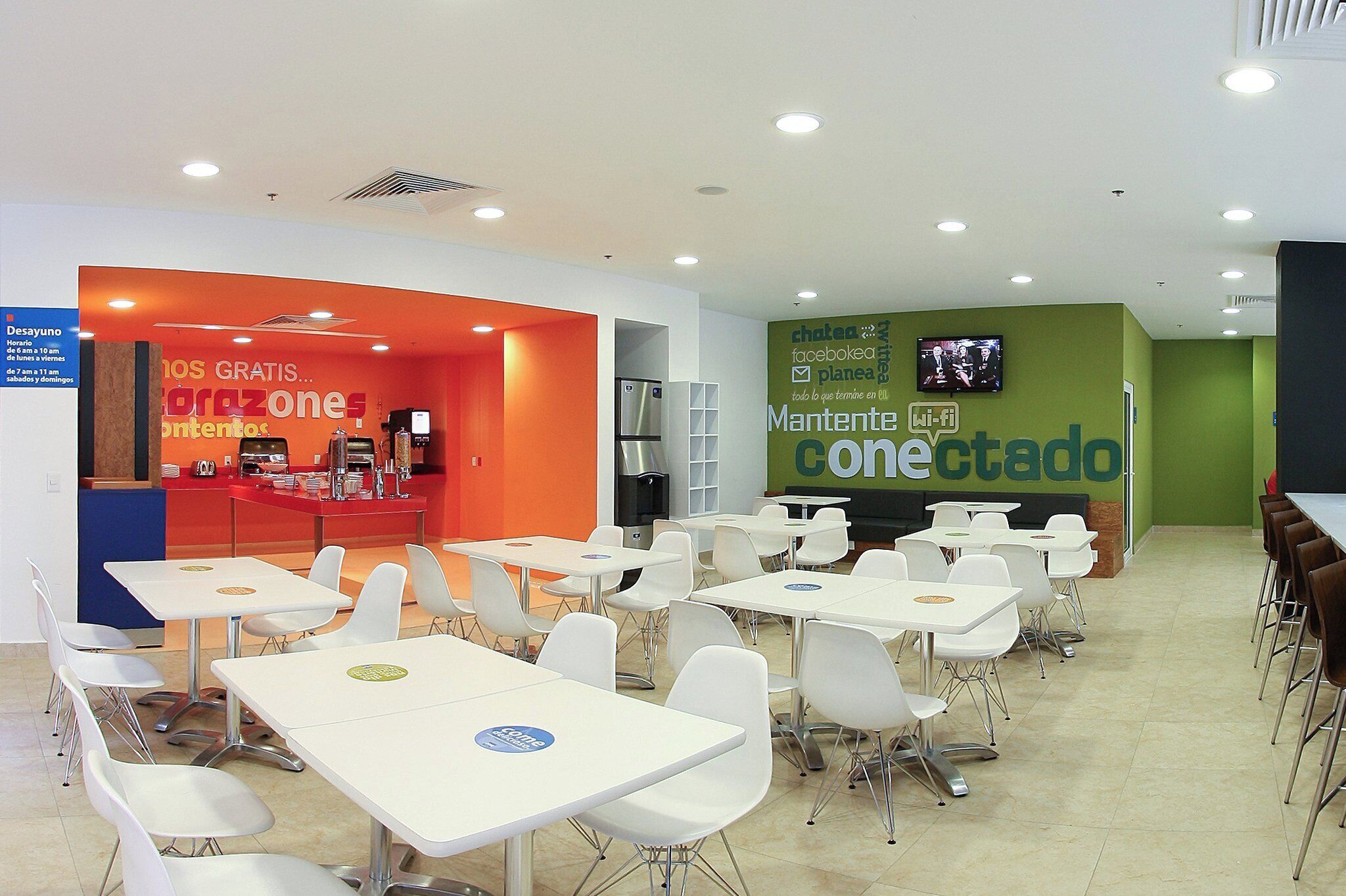 A modern cafeteria with white tables and chairs, a counter with food items, and a green wall with a Wi-Fi sign.
