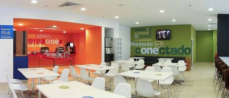 A modern cafeteria with white tables and chairs, a counter with food items, and a green wall with a Wi-Fi sign.