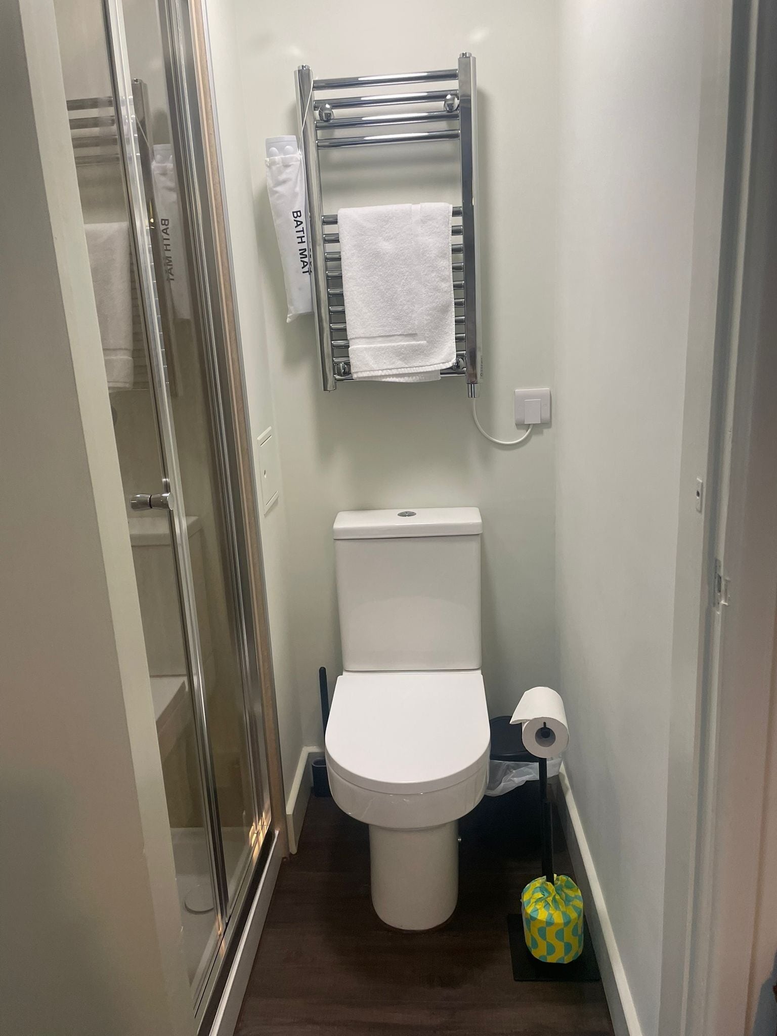 A compact bathroom with a white toilet, a towel rack with a white towel, a toilet paper holder, and a trash bin.