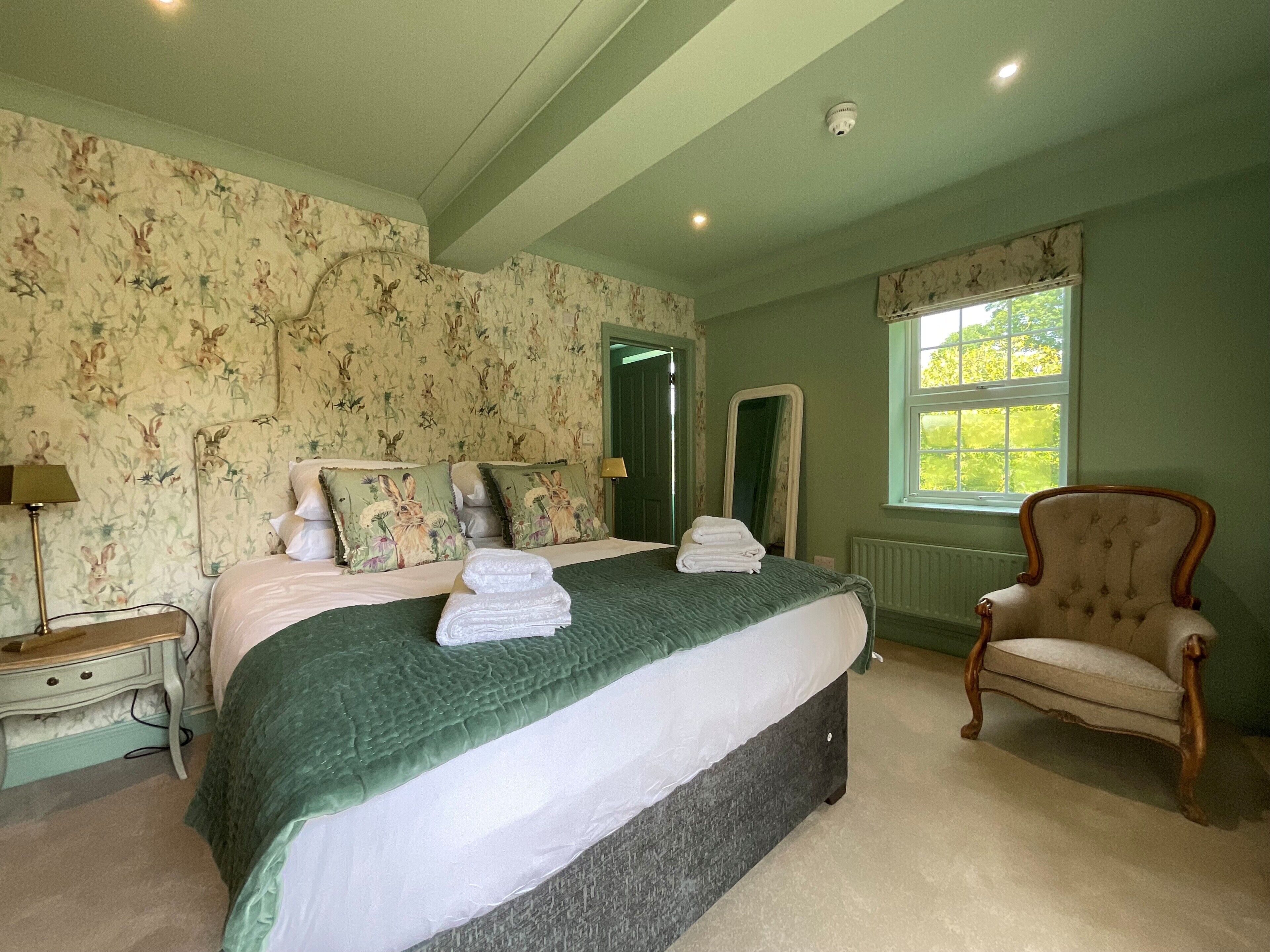 A bedroom with a large bed, a chair, a nightstand, a mirror, and a window with daylight.