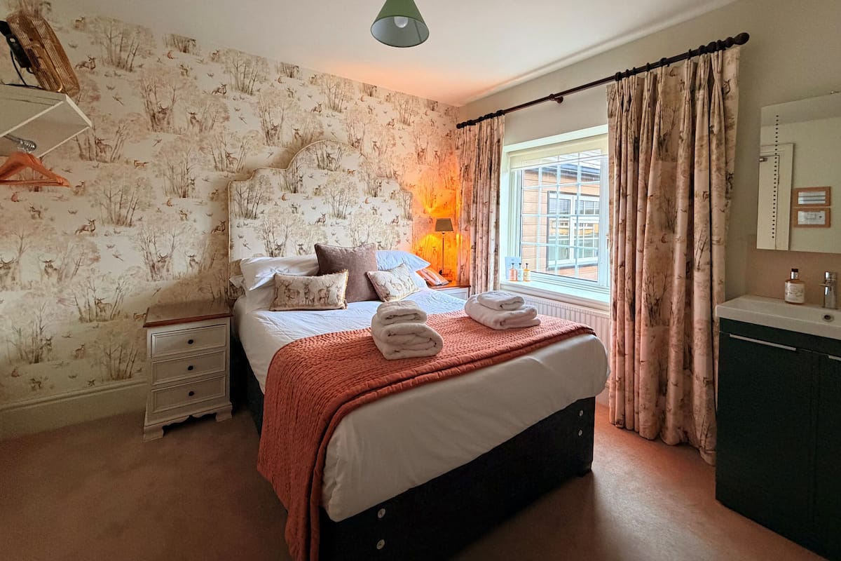 Une chambre à coucher comprenant un lit, une table de chevet, une lampe, une fenêtre avec des rideaux, une commode et un mur recouvert d’un papier peint à motifs de cerfs.