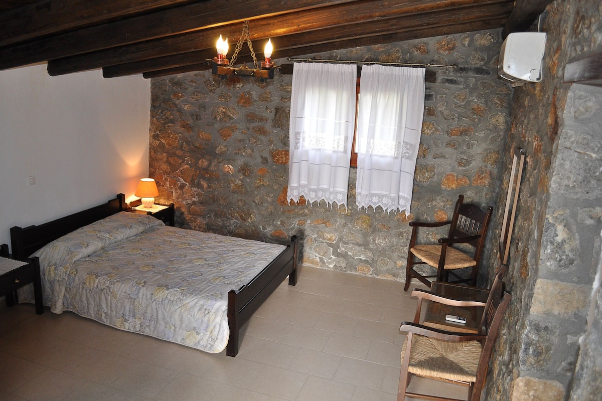 A bedroom with a bed, a chair, a nightstand, a lamp, and a stone wall.