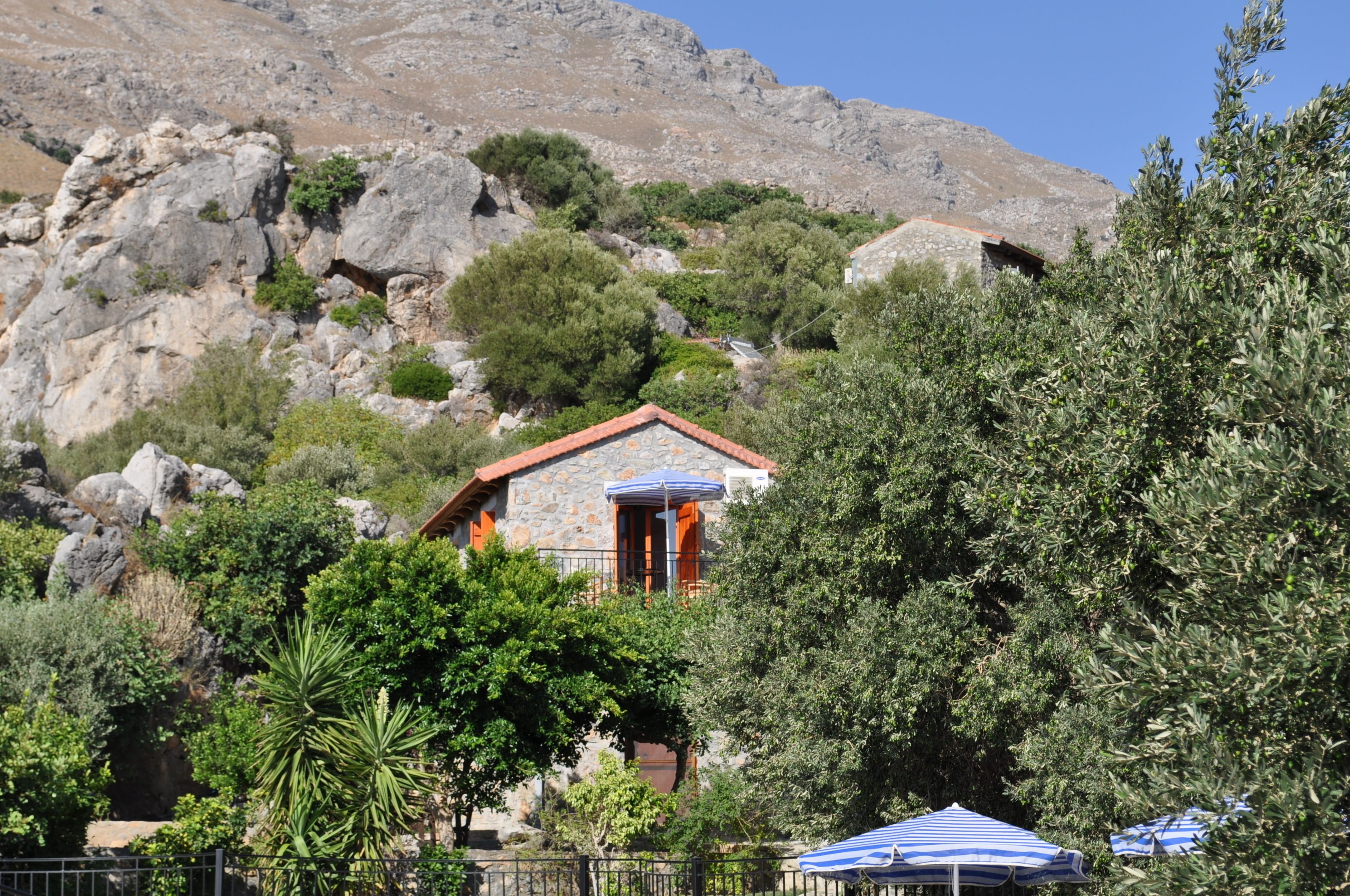 A stone building with a red-tiled roof, surrounded by lush greenery and rocky hills.