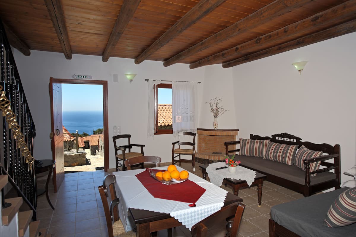 A cozy living room with a wooden ceiling, a sofa with striped cushions, a dining table with a bowl of oranges, and a view of the sea through an open door.