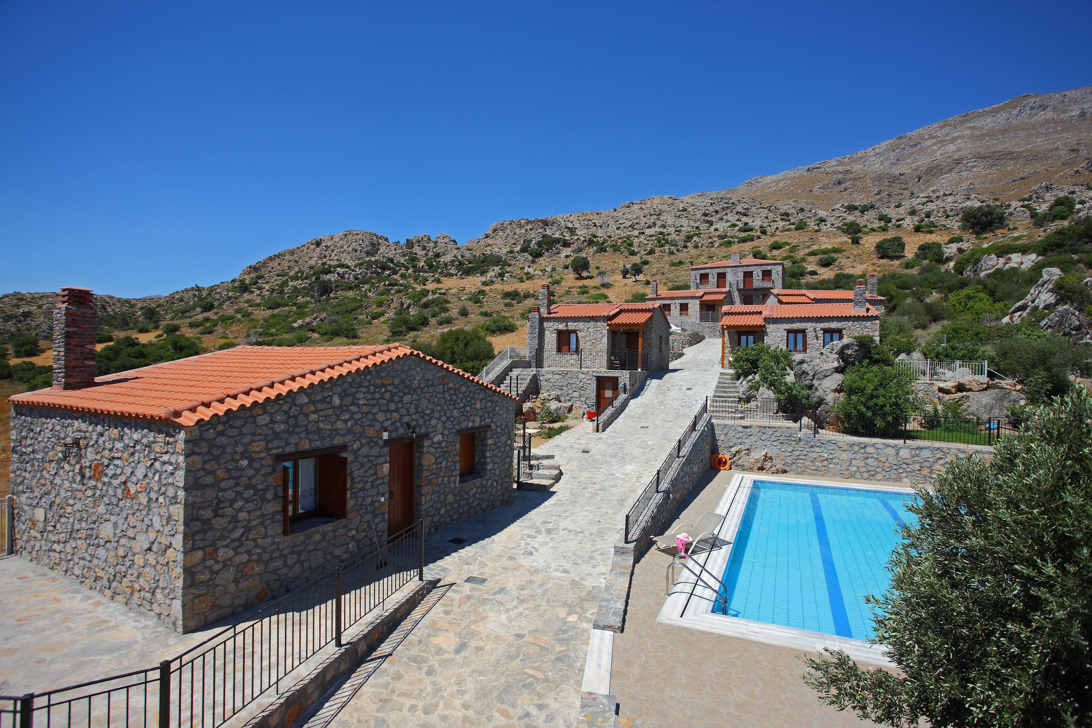 Un edificio de piedra con techo de tejas rojas, piscina y montañas al fondo.