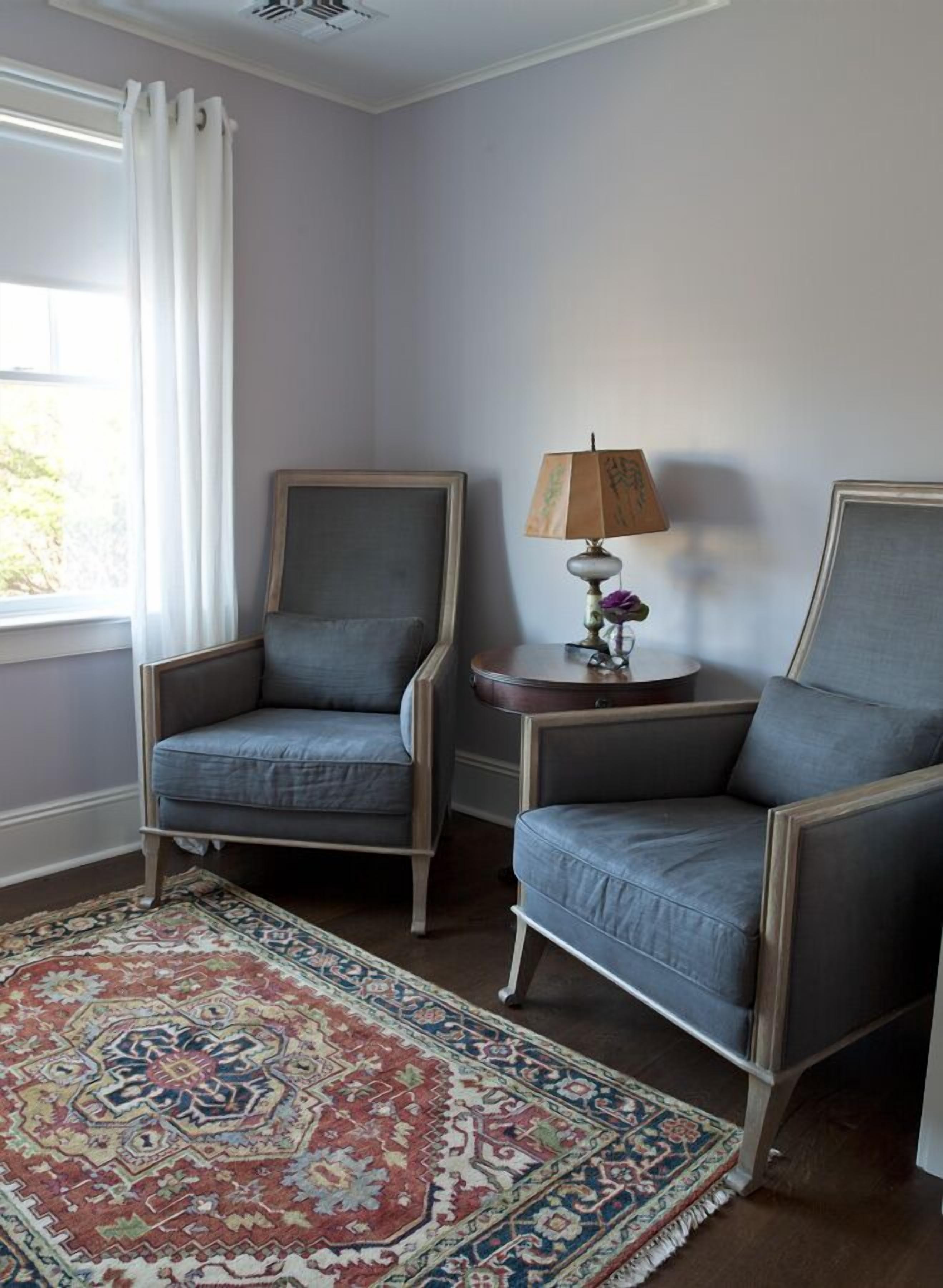 Una habitación con dos sillones grises, un tapete estampado, una lámpara con pantalla floral y una ventana con cortinas blancas.