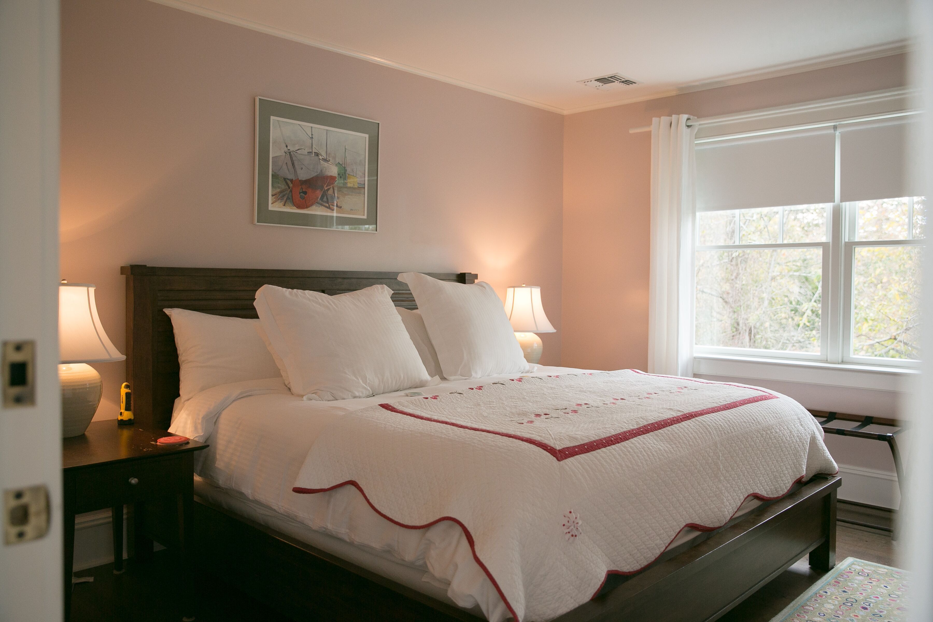 A neatly made bed with white linens and a red trim, a nightstand with a lamp, and a window with a view of trees.