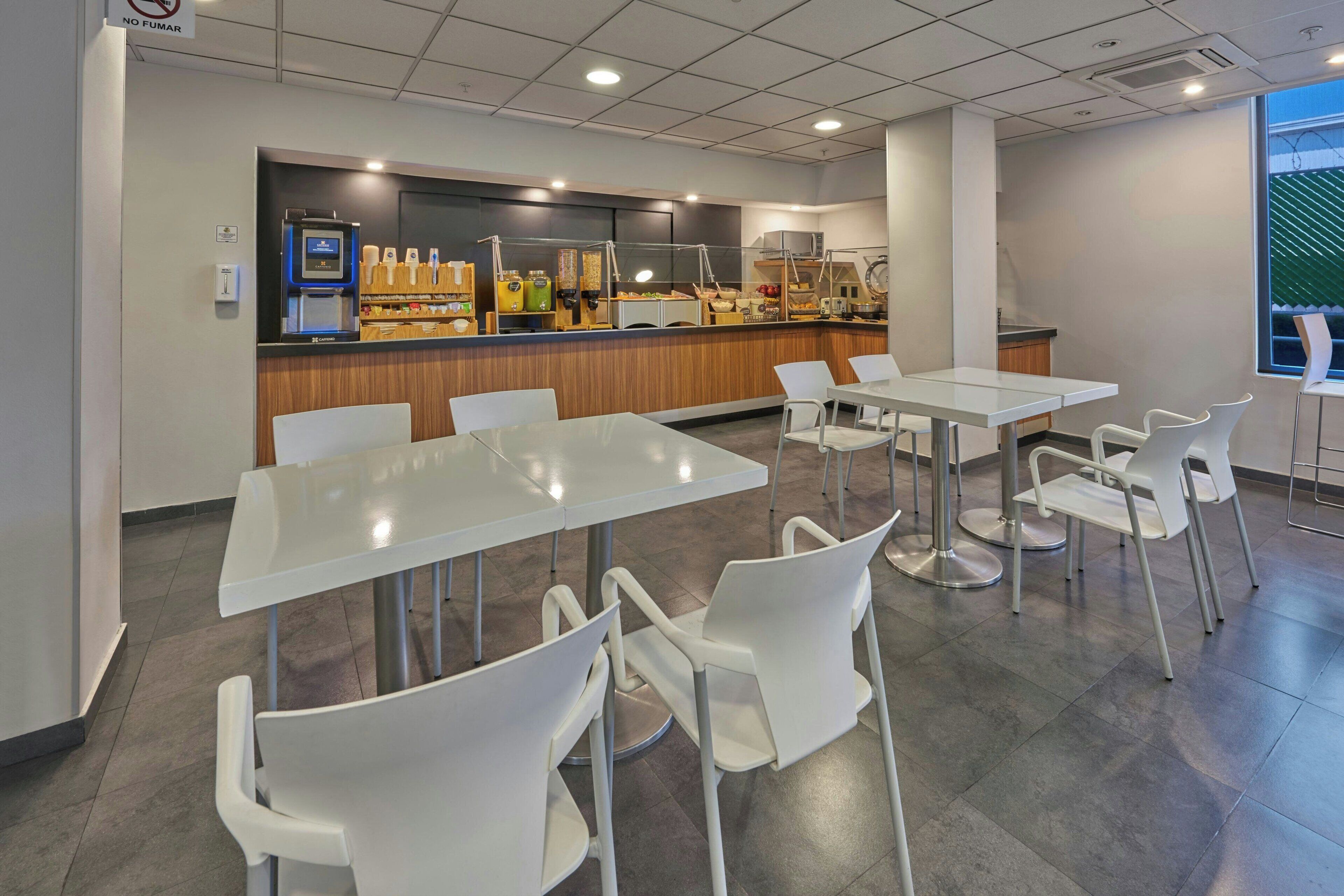 A modern cafe with white tables and chairs, a counter with various food items, and a view of the outside through a window.