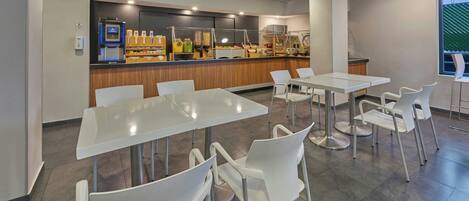 Un café moderno con mesas y sillas blancas, una barra con diversos alimentos y vistas al exterior a través de un ventanal.