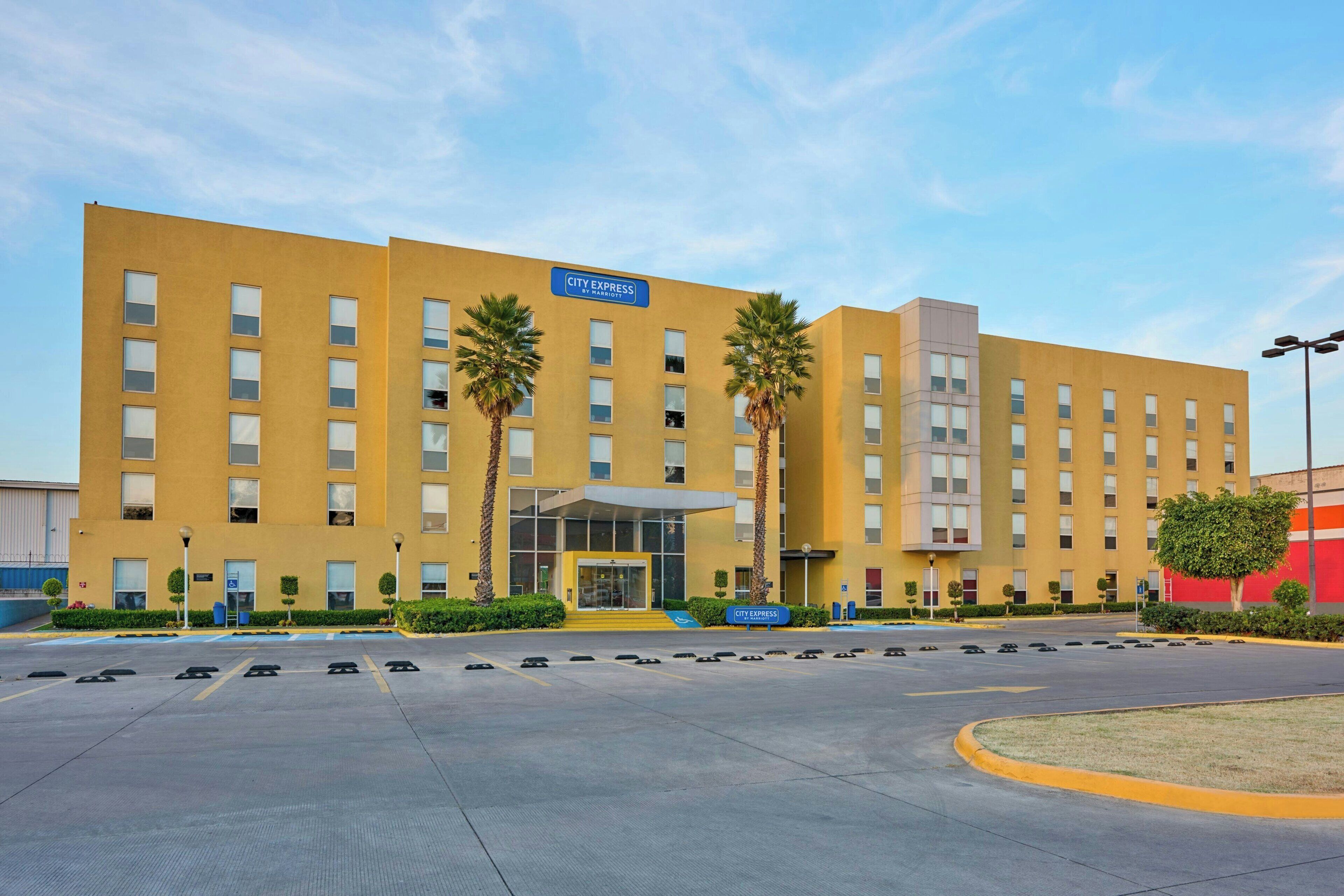 Un hotel City Express con fachada amarilla, palmeras y un estacionamiento.