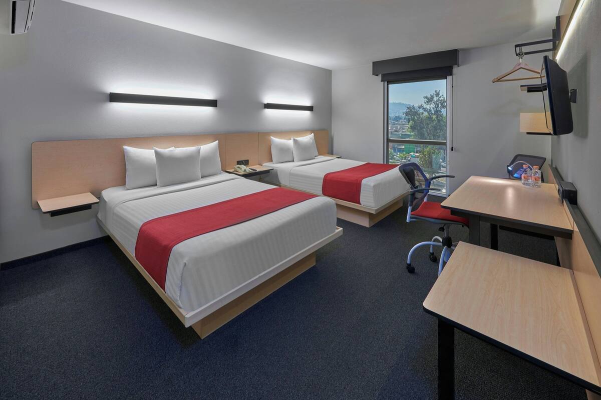Ein Hotelzimmer mit zwei Betten, einem Schreibtisch, einem Stuhl, einem Fernseher und einem Fenster mit Blick auf die Stadt.