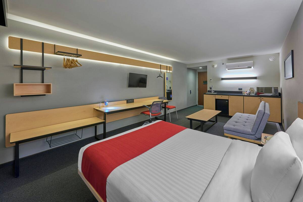 Ein modernes Hotelzimmer mit einem großen Bett, einem Schreibtisch, einem Sessel, einer Bank, einem Fernseher und einer Küchenzeile.