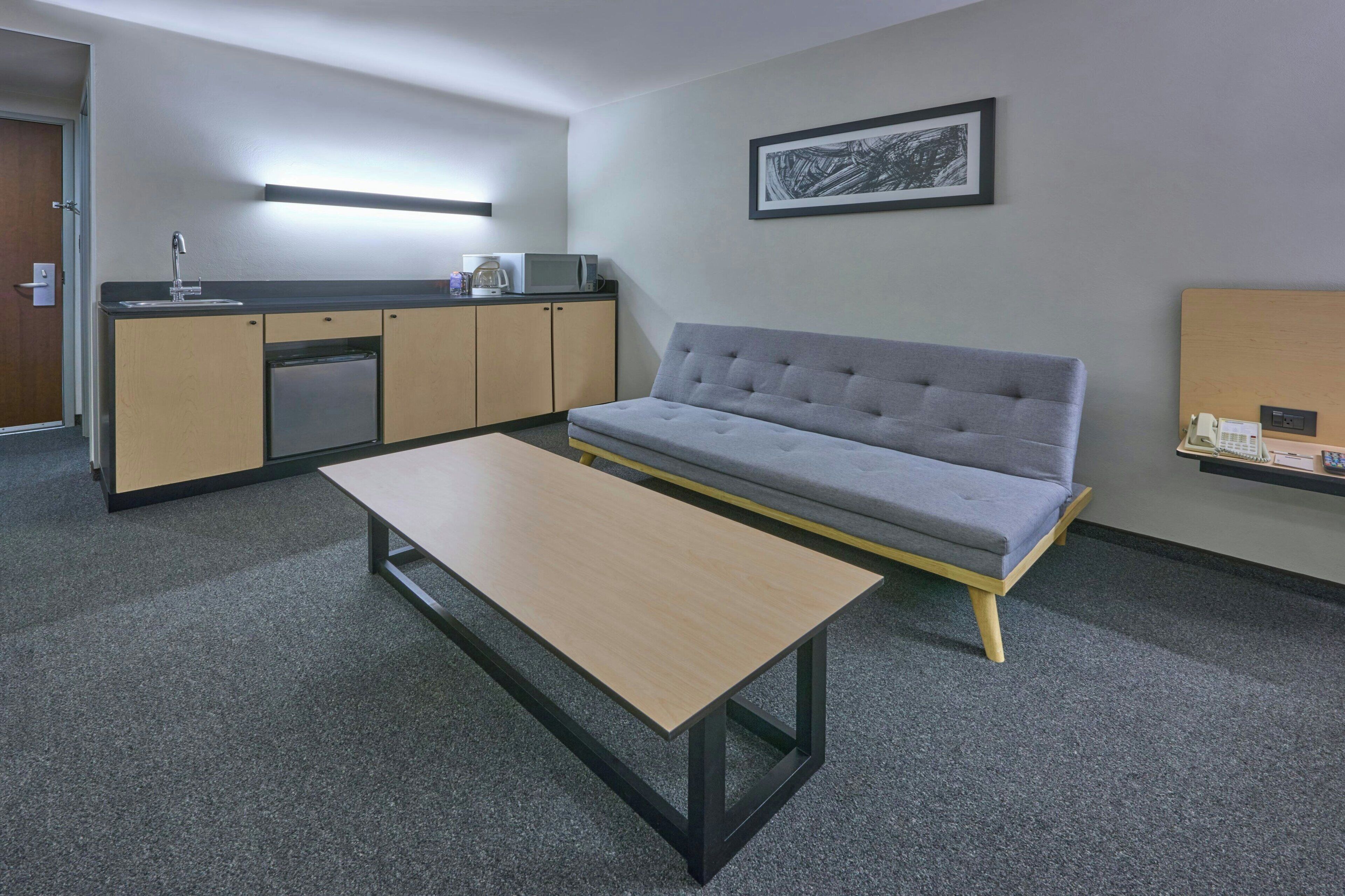 A compact hotel room with a kitchenette, a grey sofa, a small table, and a framed picture on the wall.