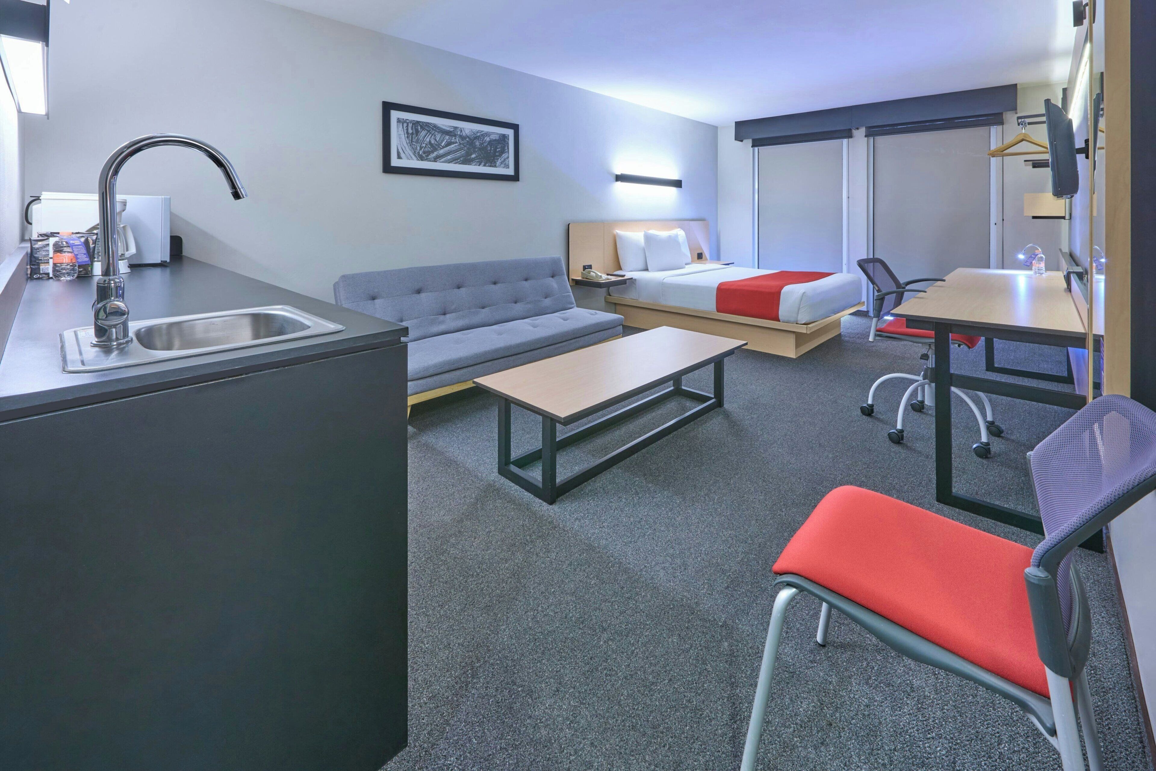 A modern hotel room with a kitchenette, a sofa, a bed with a red accent, a desk, and a chair.