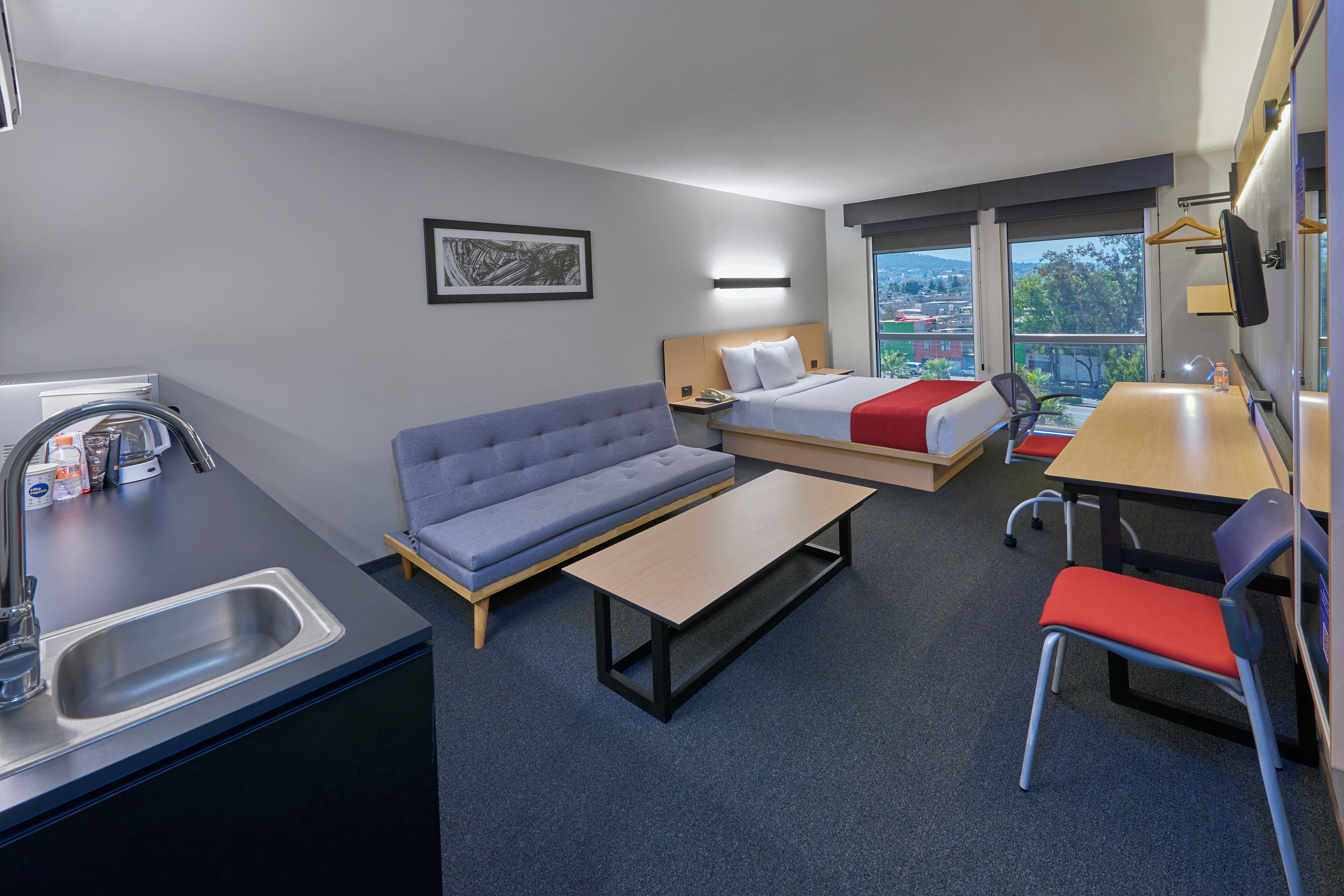 A compact hotel room with a kitchenette, a sofa, a bed with a red blanket, a desk, and a chair.