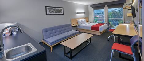 A compact hotel room with a kitchenette, a sofa, a bed with a red blanket, a desk, and a chair.