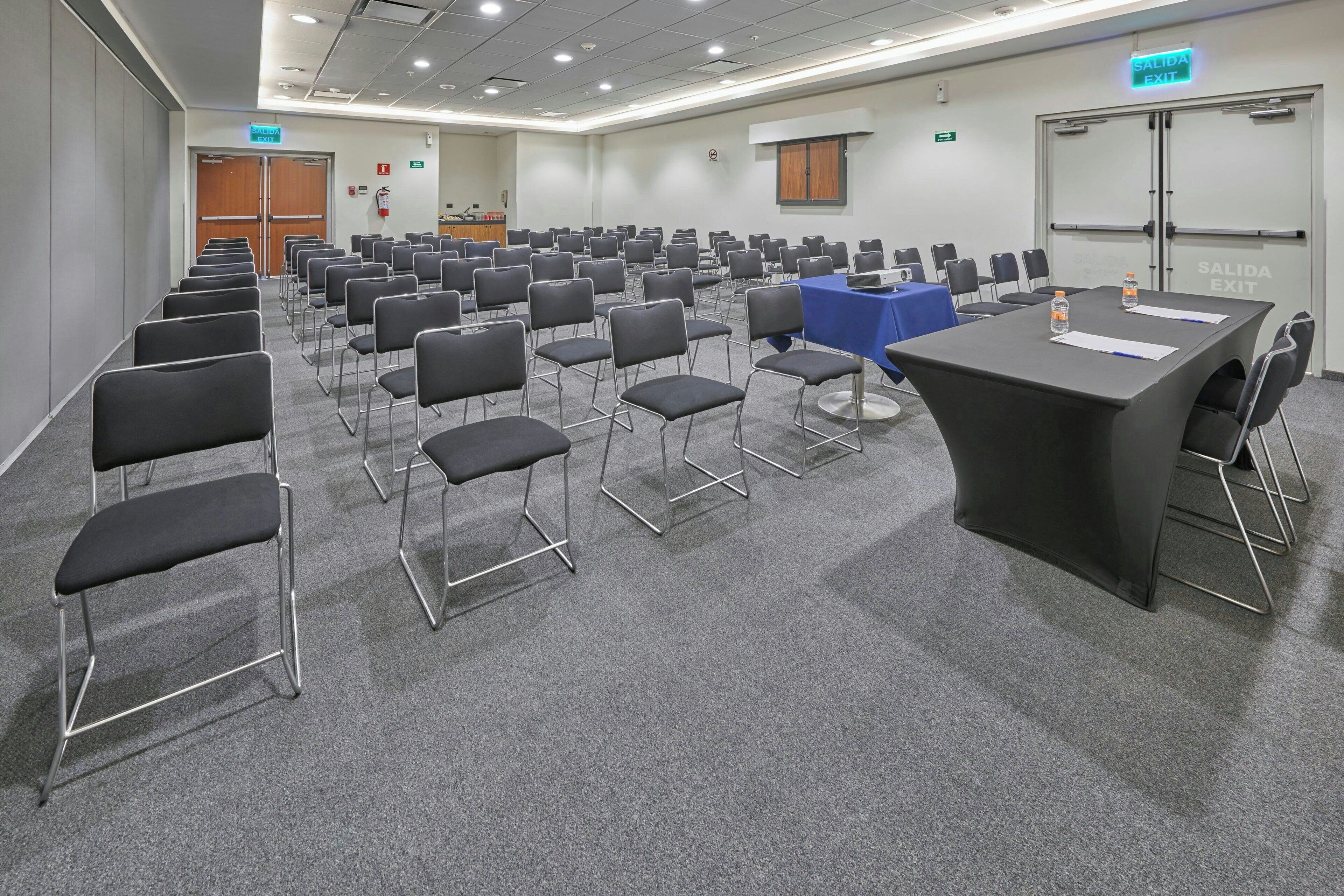 A conference room with rows of chairs, a long table with a blue tablecloth, and an exit sign.