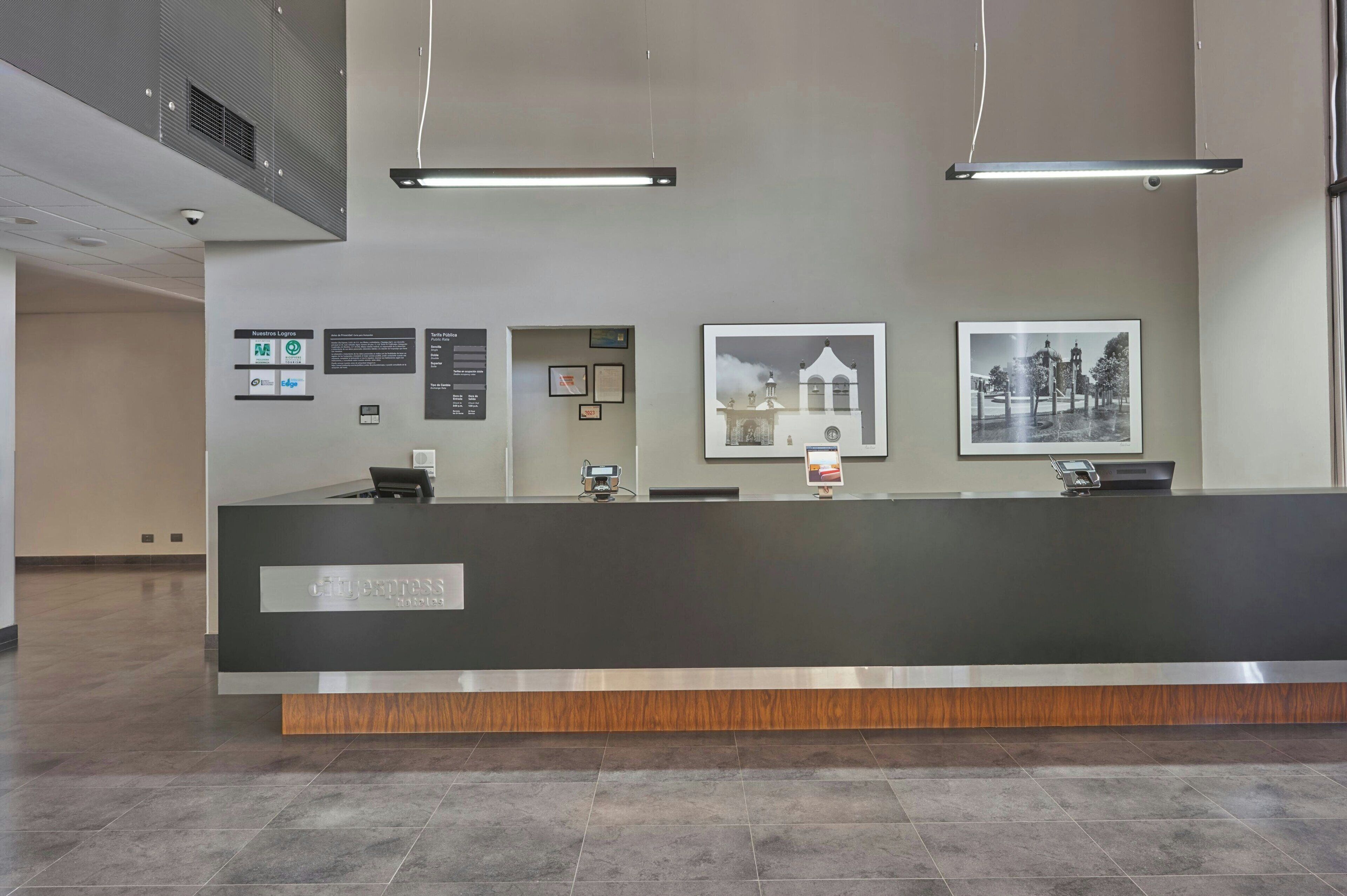 Un comptoir d’accueil avec un plan de travail foncé, un ordinateur et un téléphone. Des tableaux encadrés ornent le mur, ainsi qu’un panneau affichant des logos d’entreprises.
