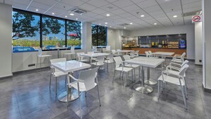 Un comedor moderno con mesas y sillas blancas, ventanas amplias con vista a coches estacionados y árboles, y una cocina con mostrador y electrodomésticos.