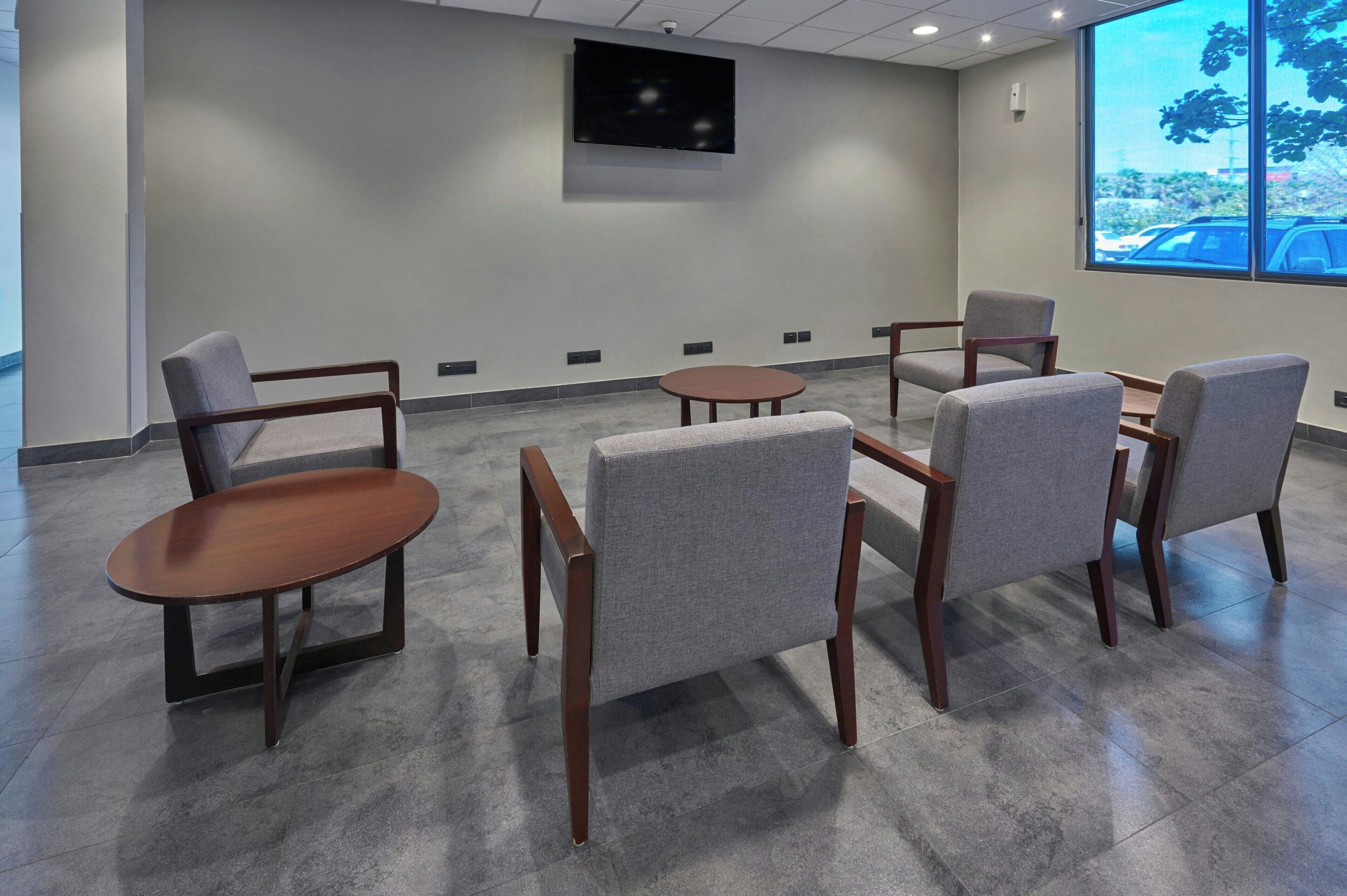 Une salle de conférence avec un grand téléviseur accroché au mur, des chaises grises, une table ronde et une vue sur un parking, visible à travers une grande fenêtre.