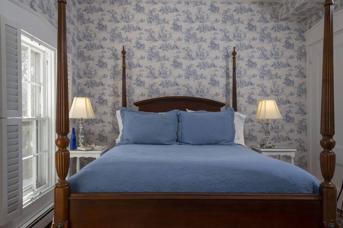 A four-poster bed with a blue bedspread, two bedside tables with lamps, and floral wallpaper.
