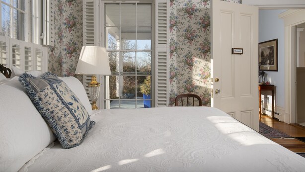 A bedroom with a large bed, floral wallpaper, a door with a nameplate, and a view through the window. - Liberty Hill Inn (Yarmouth Port)