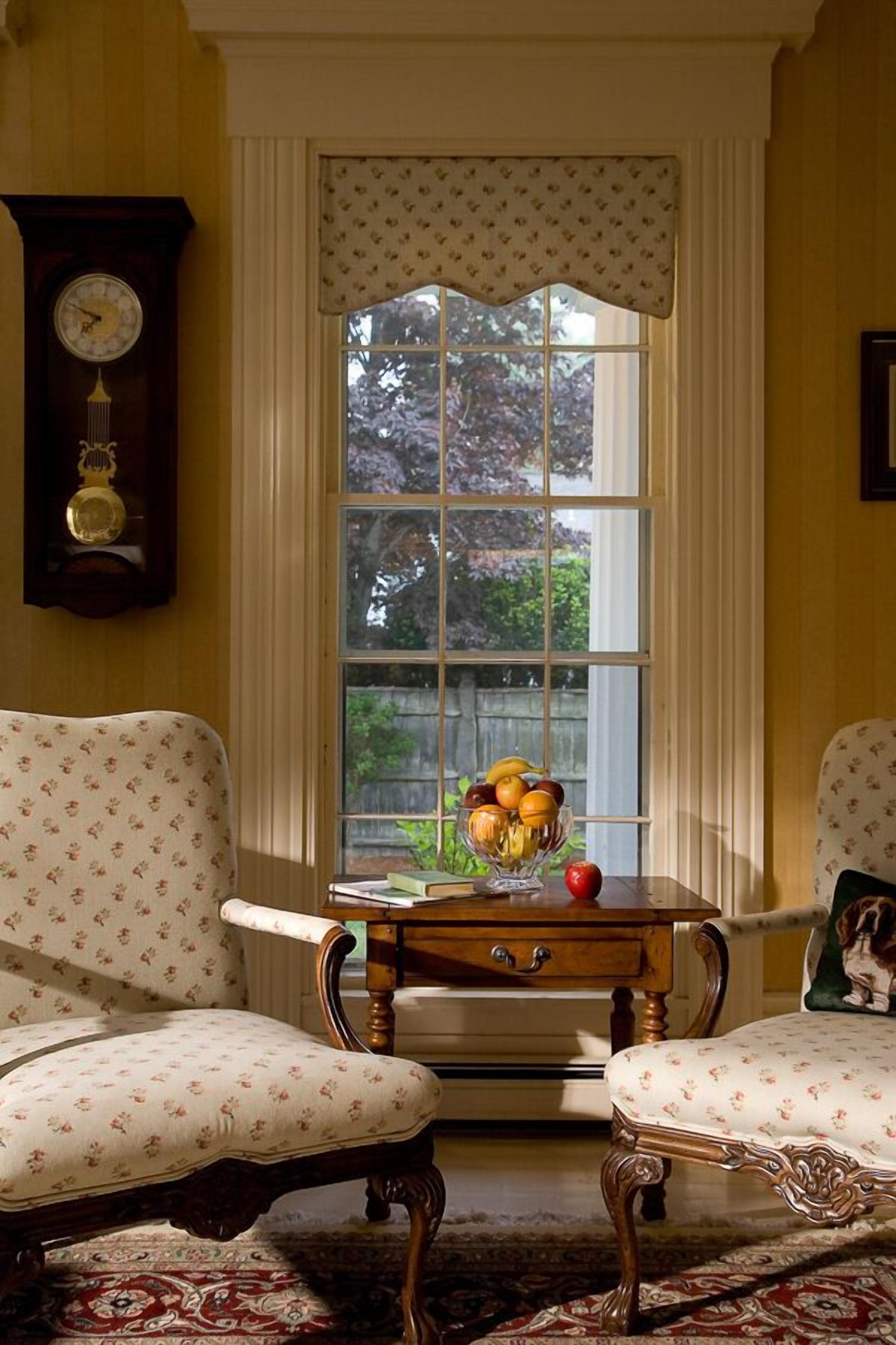 A room with two upholstered chairs, a wooden side table with a fruit bowl, a clock, and a window with curtains.