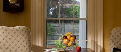 A room with two upholstered chairs, a wooden side table with a fruit bowl, a clock, and a window with curtains.