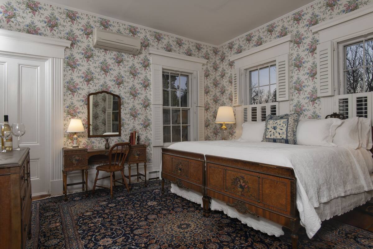A bedroom with floral wallpaper, a wooden bed, a desk with a chair, and a large window with shutters.