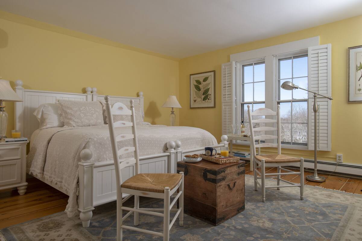 A bedroom with a bed, two bedside tables, a dresser, a chair, and a window with shutters.