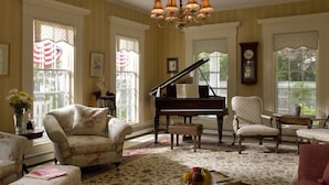 A room with a grand piano, floral armchairs, and a chandelier. - Liberty Hill Inn (Yarmouth Port)