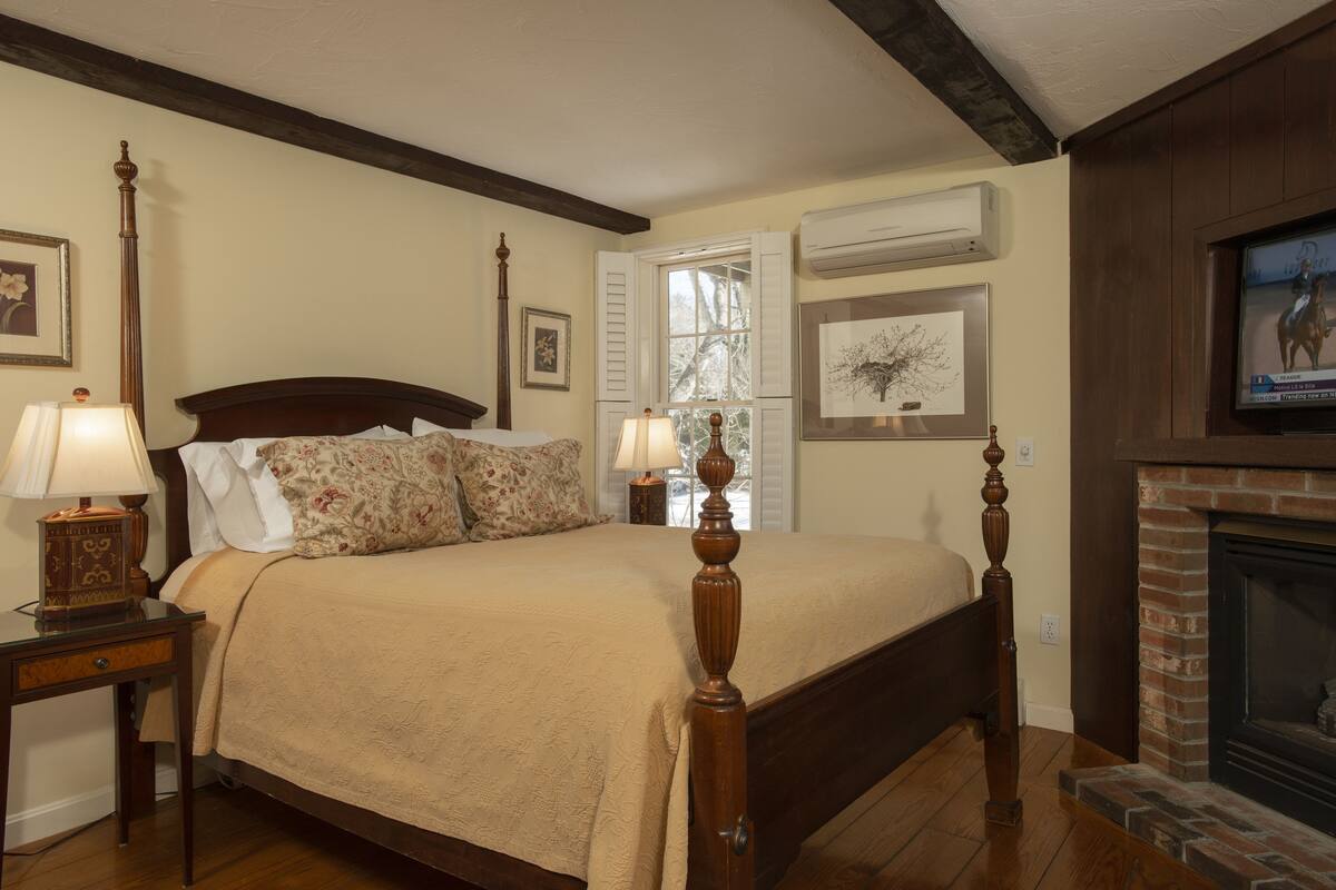 A bedroom with a large bed, wooden headboard, two bedside tables with lamps, a fireplace, and framed artwork on the walls.