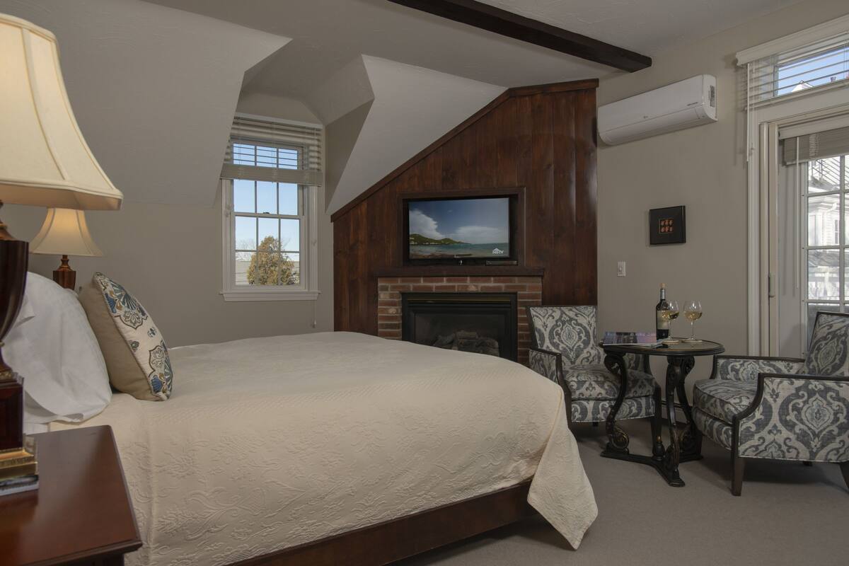 A bedroom with a large bed, a fireplace, a TV mounted above it, and two patterned armchairs.