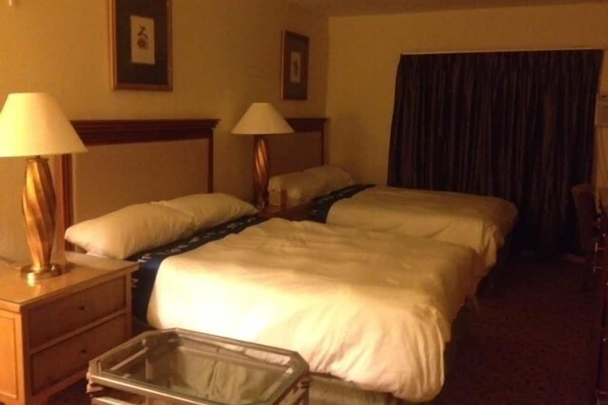 A hotel room with two beds, a nightstand, a lamp, and a small table.