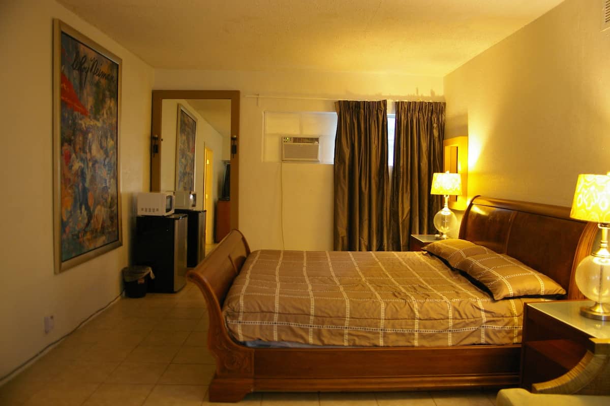 A hotel room with a bed, a nightstand, a lamp, a painting on the wall, and an air conditioning unit.