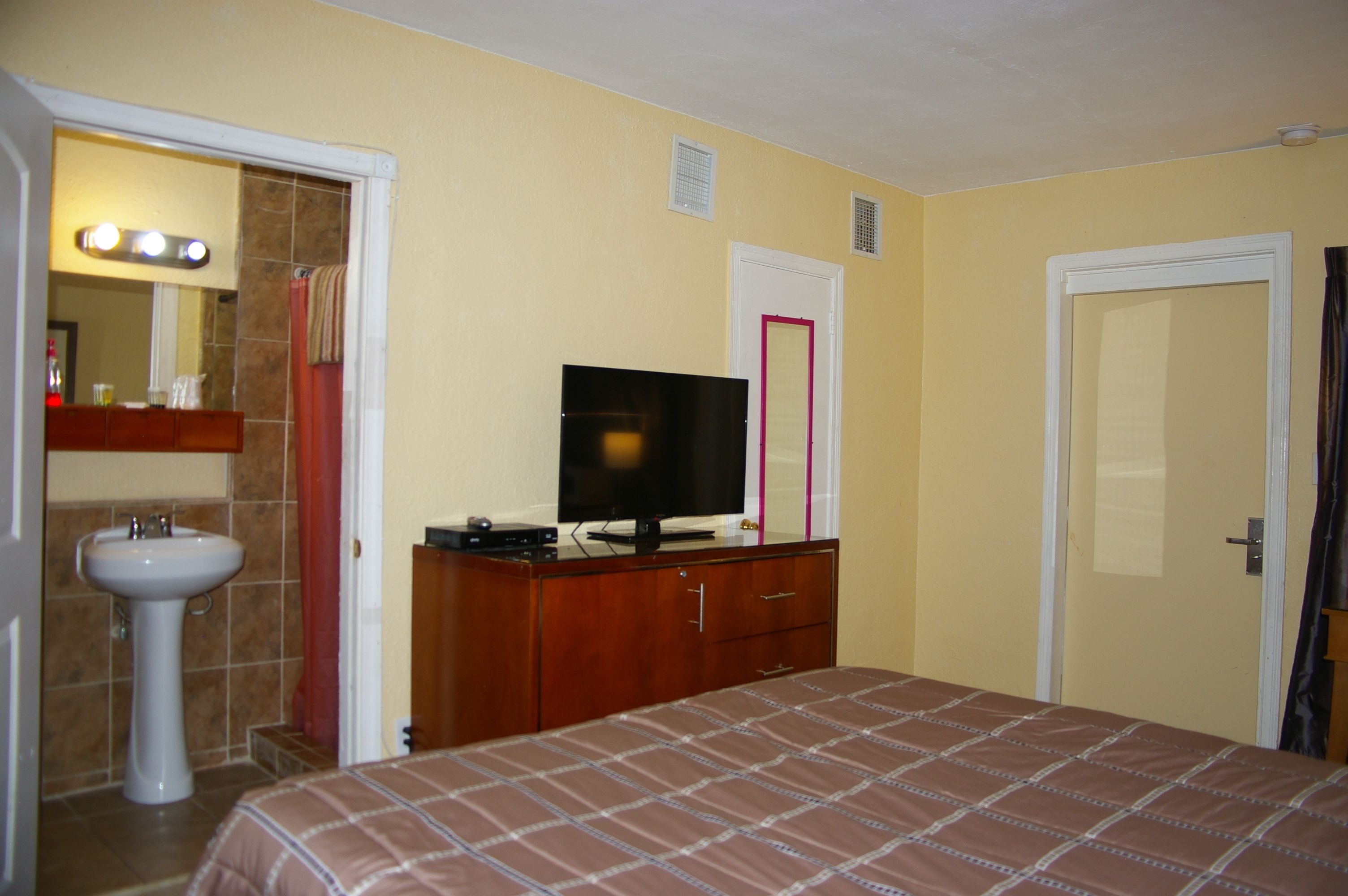 Una habitación de hotel con cama, televisión sobre un aparador de madera, lavamanos y espejo con luces.