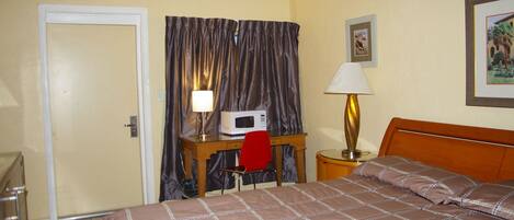 Habitación de hotel con cama, escritorio, silla, microondas, lámpara y puerta.