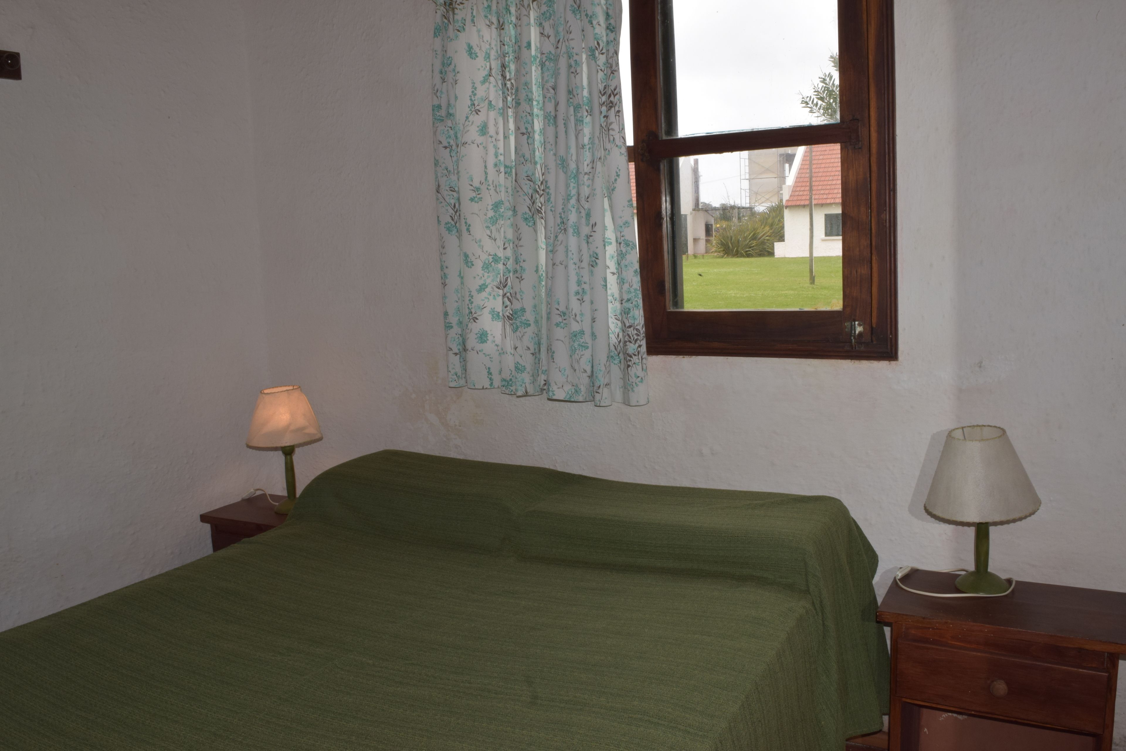 A room with a bed, a window with curtains, a nightstand with a lamp, and a view of a house outside.