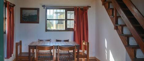 Un comedor con una mesa y sillas de madera, una ventana con cortinas rojas, una escalera de madera y una lámpara de techo tejida.