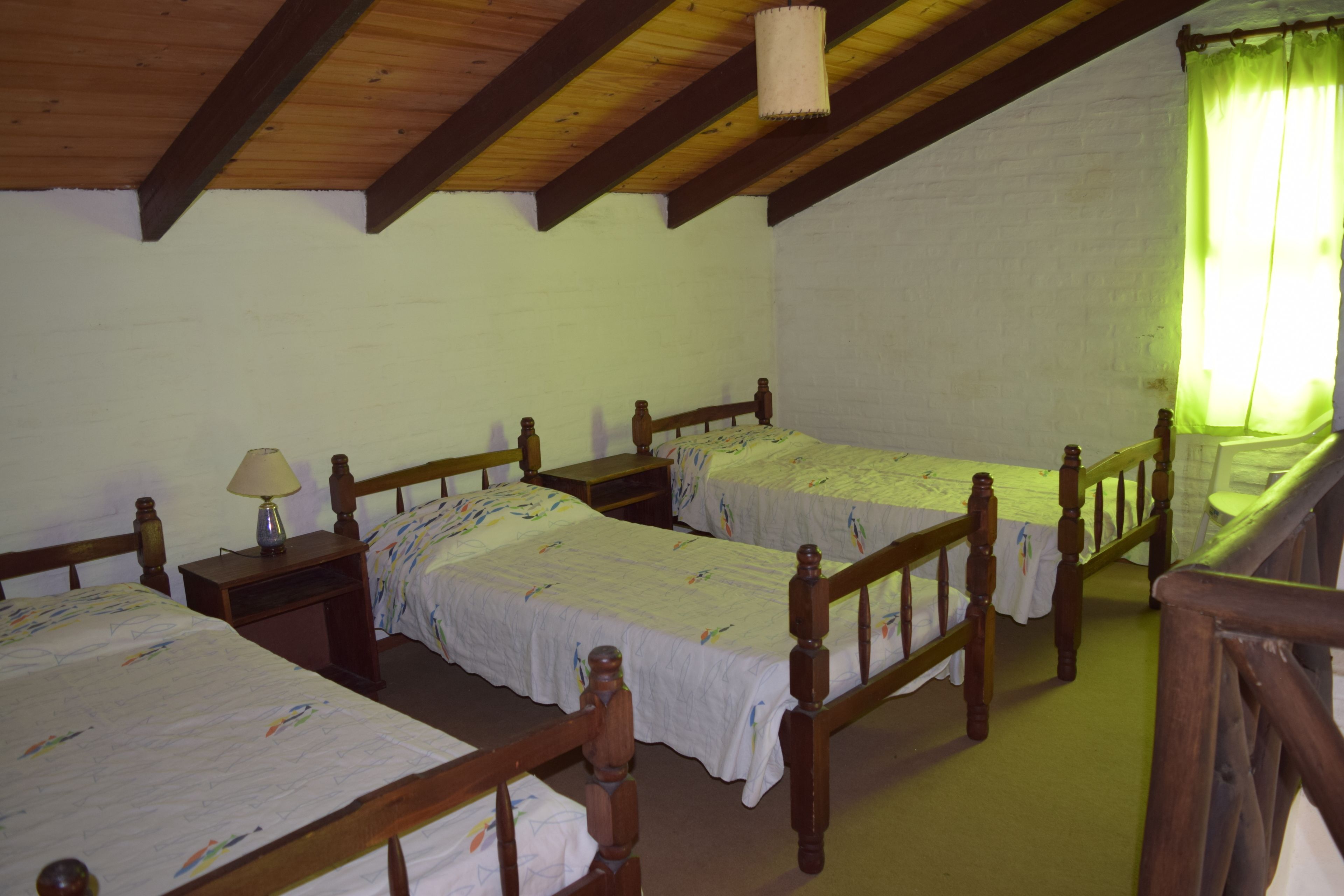Una habitación con dos camas de madera, cada una con cabecero de madera y una mesita de noche con lámpara. Paredes blancas y techo de madera con vigas a la vista.