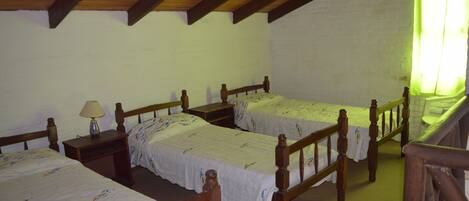 Una habitación con dos camas de madera, cada una con cabecero de madera y una mesita de noche con lámpara. Paredes blancas y techo de madera con vigas a la vista.