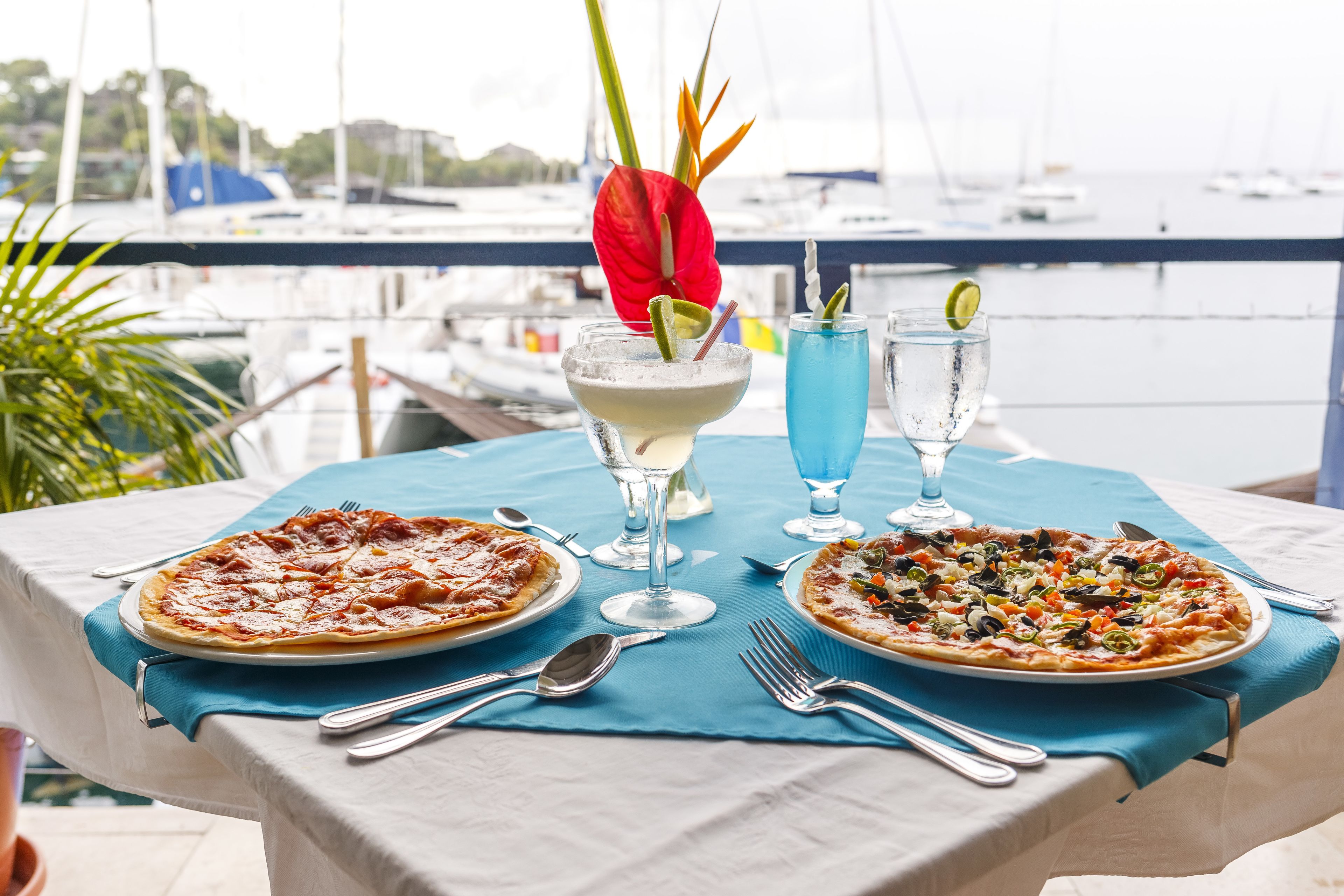Una mesa con dos pizzas, un cóctel y un vaso de agua, ubicada en un balcón con vista al puerto deportivo.