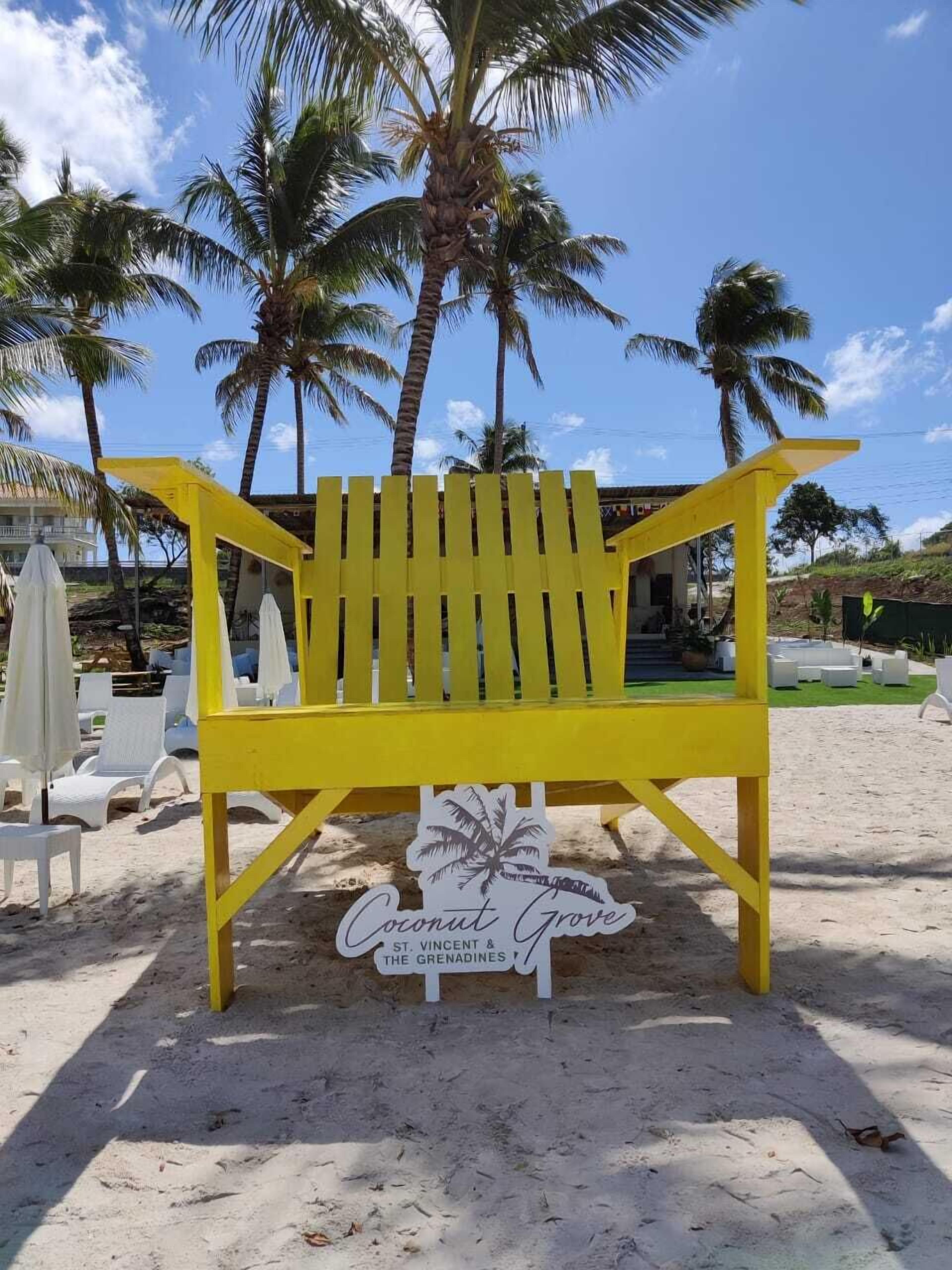 A yellow beach chair with a sign reading "Coconut Grove St. Vincent & The Grenadines" on a sandy beach with palm trees in the background.