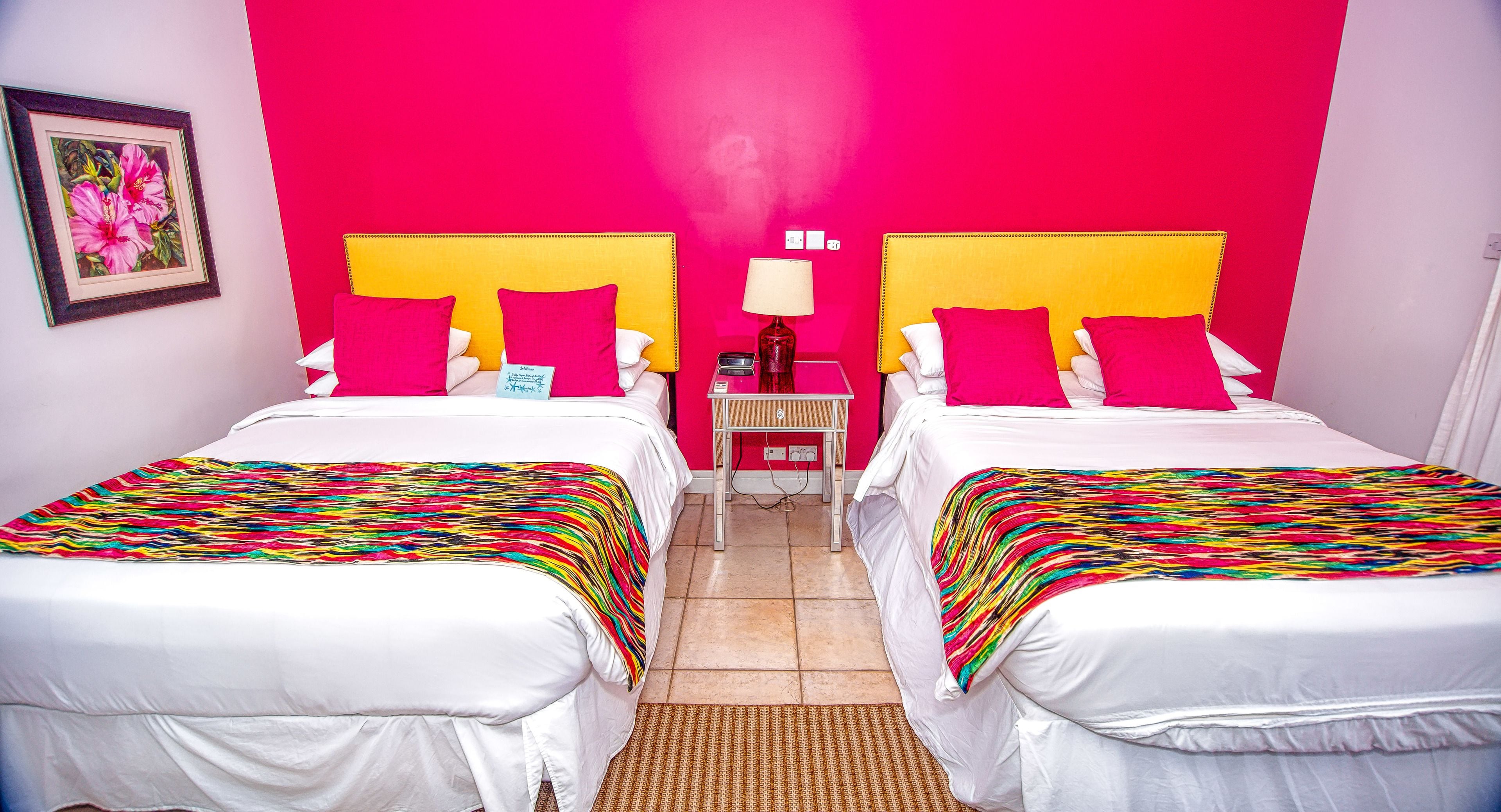 Una habitació d'hotel amb dos llits, cadascun amb un capçal groc, roba de llit blanca i llençols amb dibuixos de ratlles de colors. Hi ha una taula petita amb una làmpada i un quadre al mur.