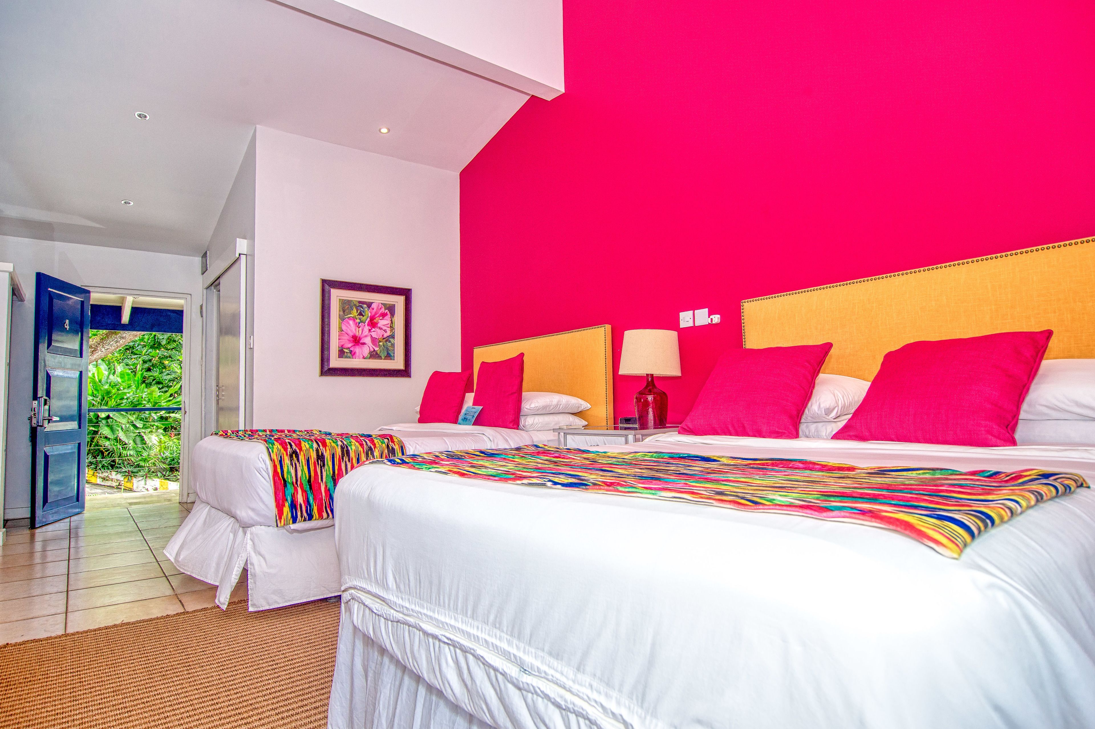 A hotel room with two beds, a bright red accent wall, and a view of greenery through an open door.